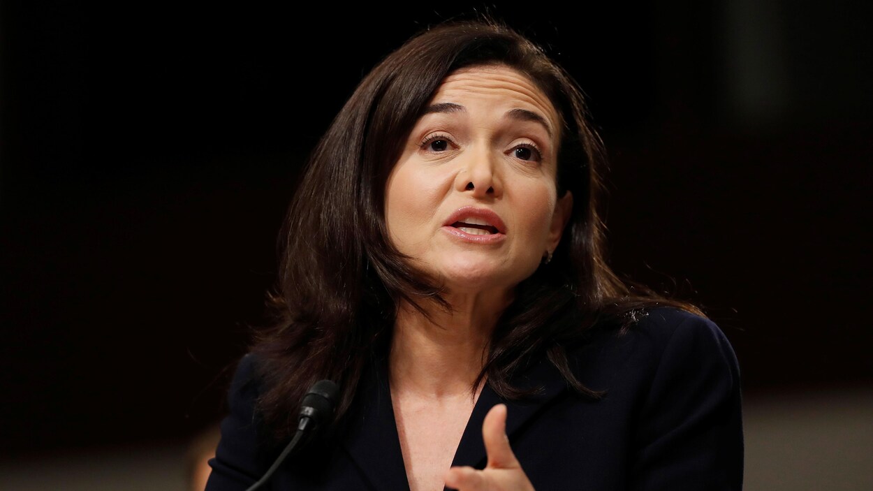 Une photo de Sheryl Sandberg portant un tailleur noir en train de parler devant un micro.