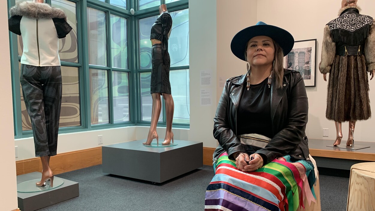 L'artiste et designer Sho Sho Esquiro assise avec en arrière-plan des mannequins vêtus de robes.