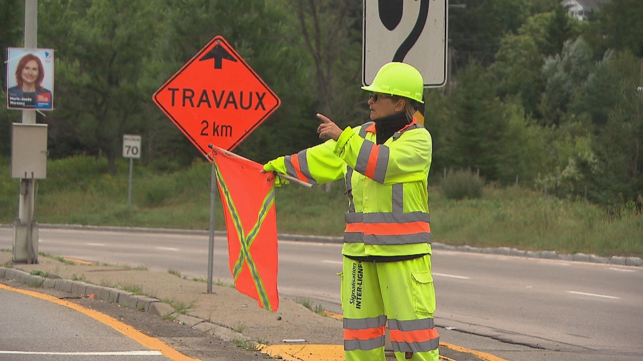 Une femme en habit de sécurité tient un drapeau orange et donne des indications avec sa main.