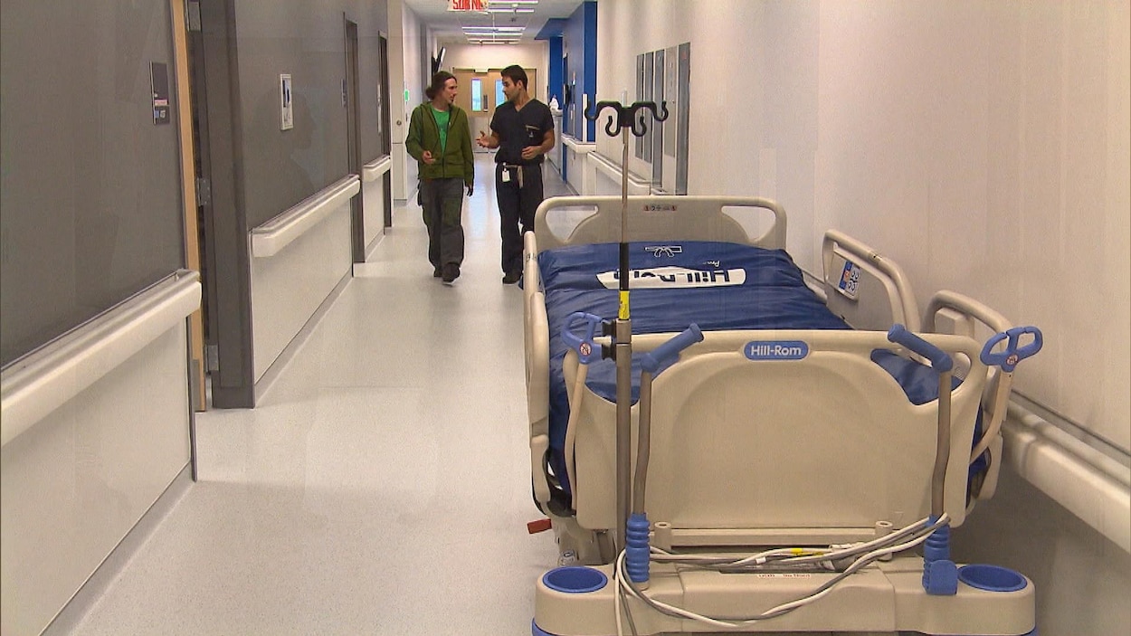 Un patient et son médecin déambulent dans le couloir d'un hôpital où l'on peut voir une civière, vide.