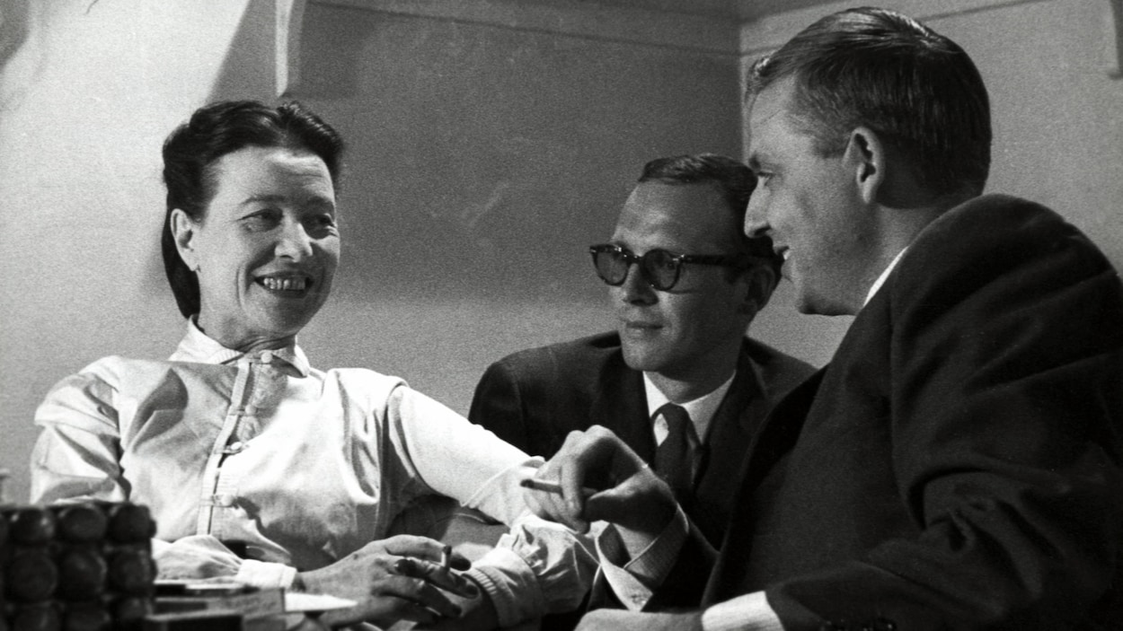Simone de Beauvoir et Wilfrid Lemoine fumant une cigarette à une petite table. Claude Sylvestre est également assis avec eux, un peu en arrière-plan.