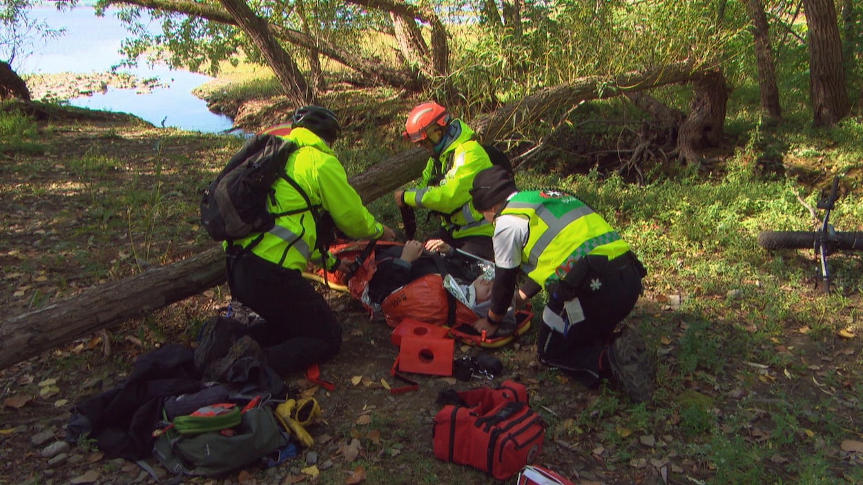 Trois personnes portant des gilets jaunes installent une femme sur une civière en bordure d'une rivière. Leur matériel jonche le sol.