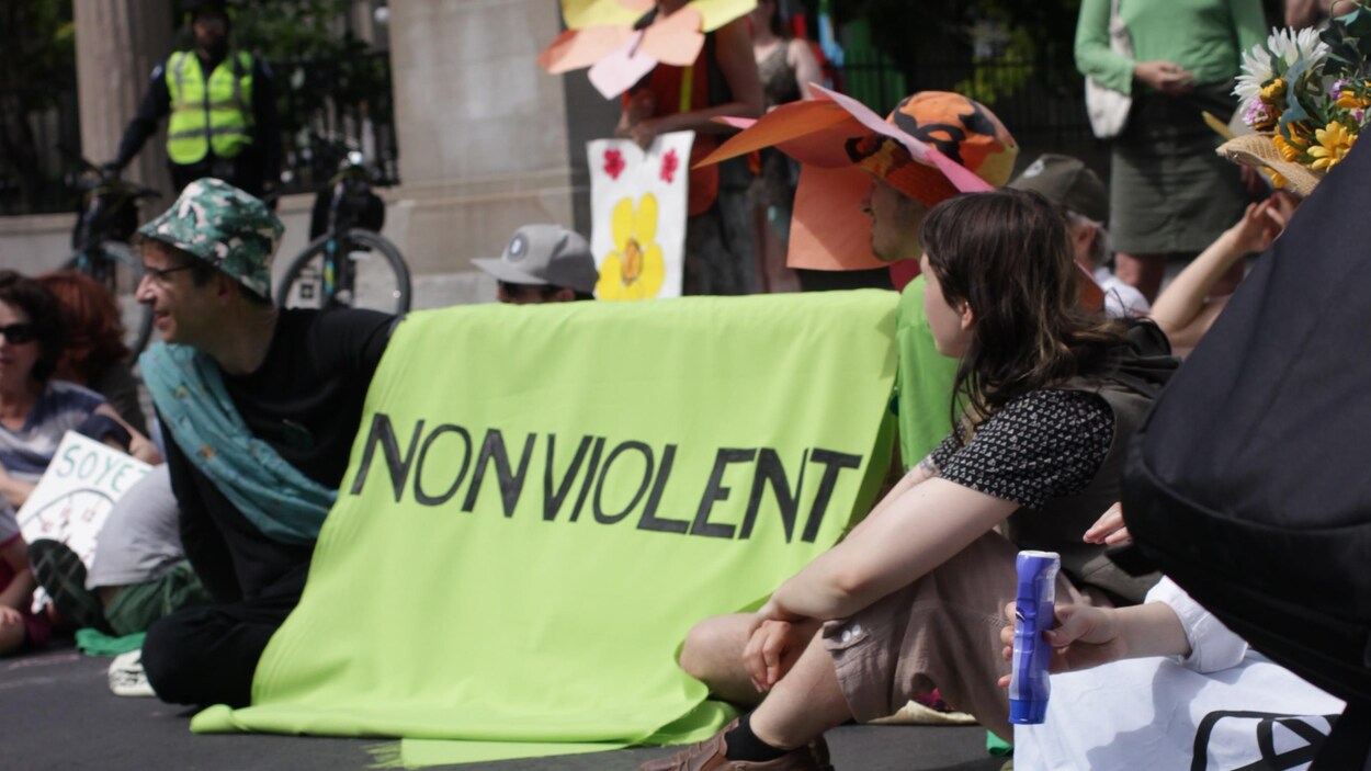 Des gens sont assis dans la rue, ils tiennent une banderole sur laquelle sont inscrits les mots « non violent » et ils portent des chapeaux ornés de fleurs.