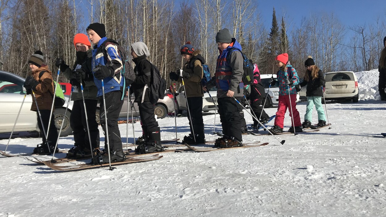 Un groupe d'élèves debout sur un terrain enneigé plat. Ils portent des ski raquettes et tiennent des bâtons de ski.