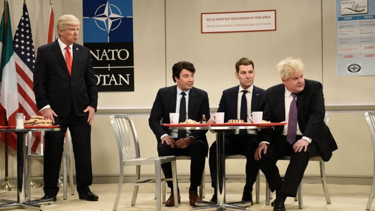 Alec Baldwin est debout et les autres sont assis autour d'une table.