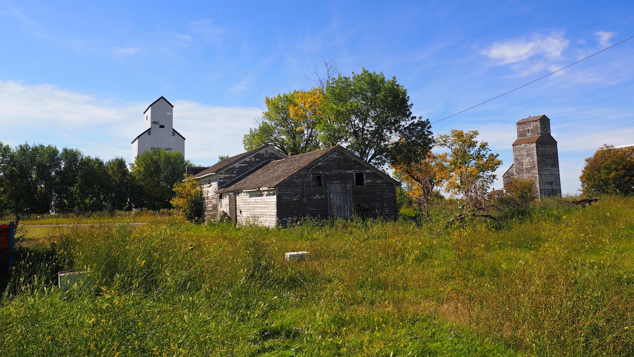 Une maison de bois abandonnée derrière laquelle on peut voir deux anciens élévateurs à grain.