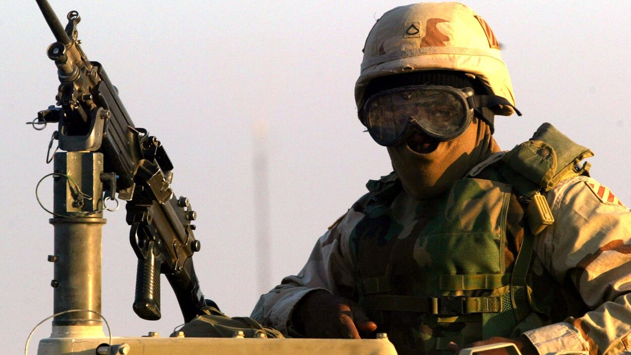 Un soldat américain portant casque, cagoule et lunettes se tient sur un véhicule d'assaut, devant une mitraillette.