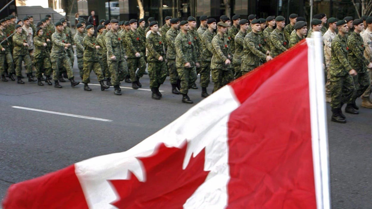 Des militaires portant l'uniforme des Forces armées canadiennes défilent à proximité d'un drapeau du Canada. Des militaires portant l'uniforme des Forces armées canadiennes défilent à proximité d'un drapeau du Canada.