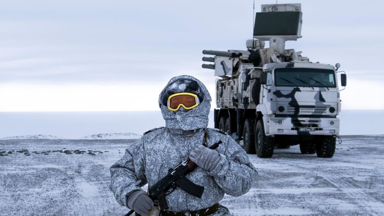 Le soldat est habillé chaudement. On ne voit que ses lunettes de ski dépasser de son capuchon. Il tient un fusil contre lui.