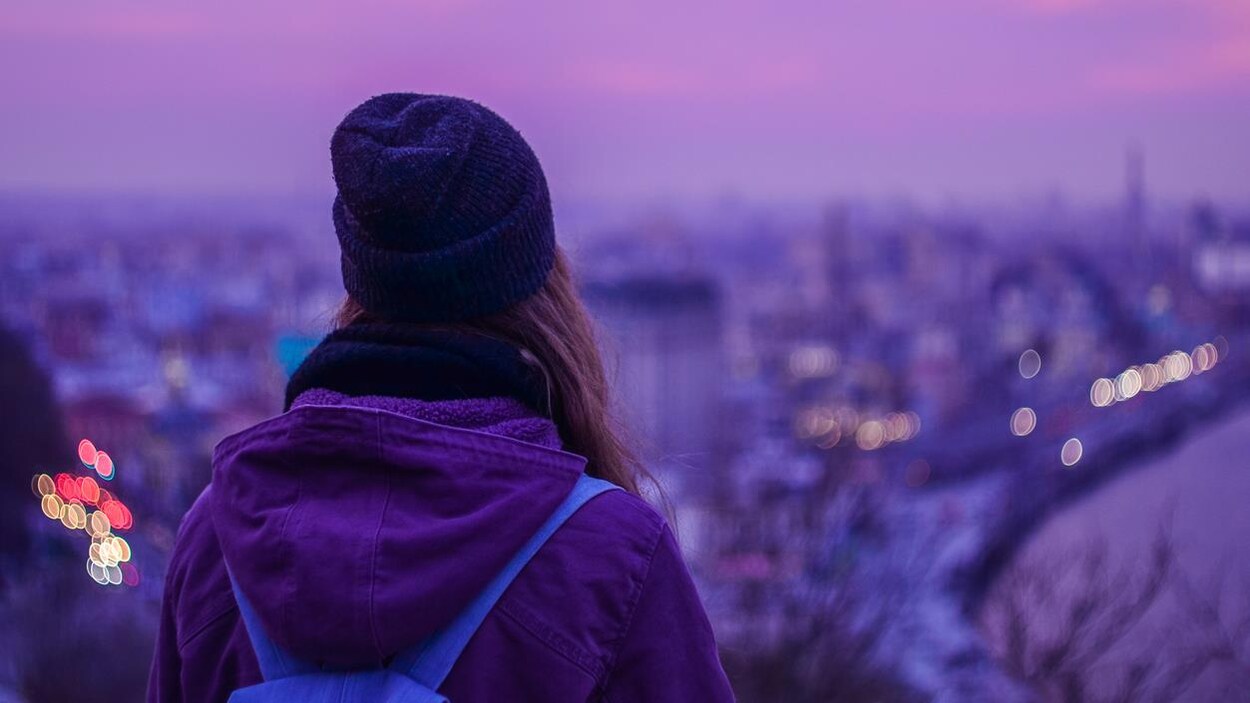 Une jeune femme portant une tuque est photographiée de dos regardant une ville au loin.