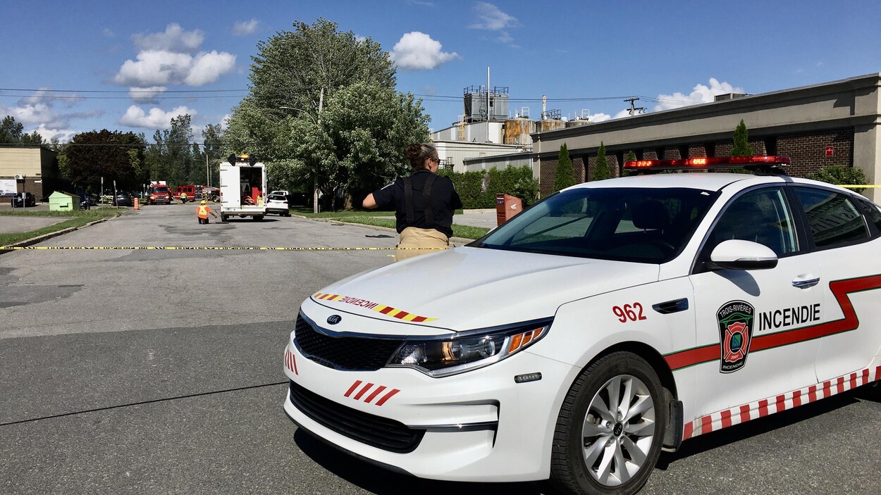 Une voiture des services incendie avec un pompier devant et un périmètre de sécurité à l'arrière-plan.
