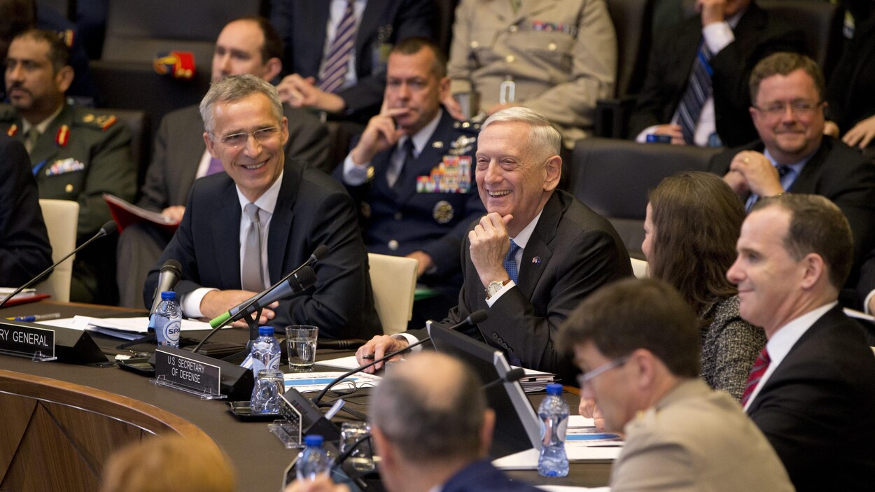 Les deux hommes assis à une table et entourés de plusieurs personnes, sourient.