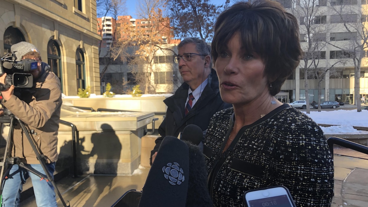 Vue sur une dame qui est debout à l'extérieur. Elle fait face à un immeuble du centre-ville de Calgary. Dans le coin gauche de la photo, il y a une caméra. En avant de la femme, il y a des micros de journalistes.