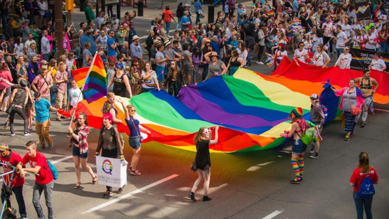 Des gens se promènent avec un immense drapeau arc-en-ciel devant une foule.