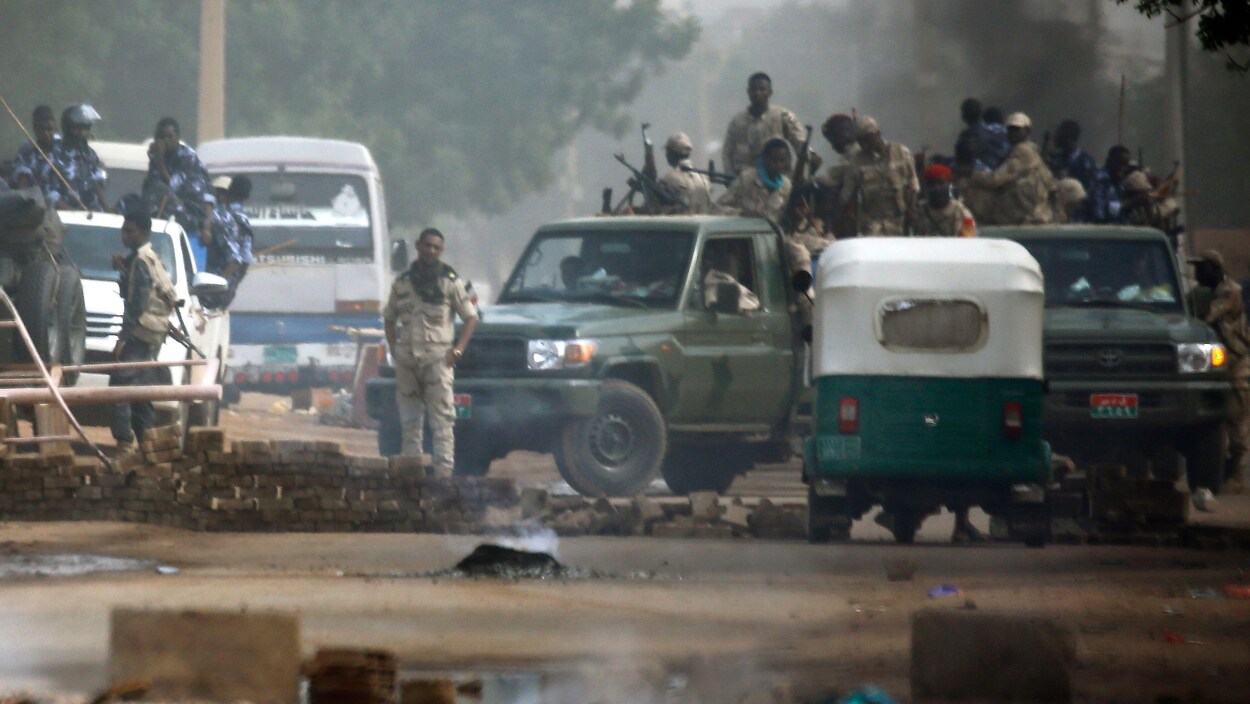 Des militaires armés, au sol ou entassés dans des boîtes de camions, sont visibles au loin, devant une barricade de fortune des manifestants.