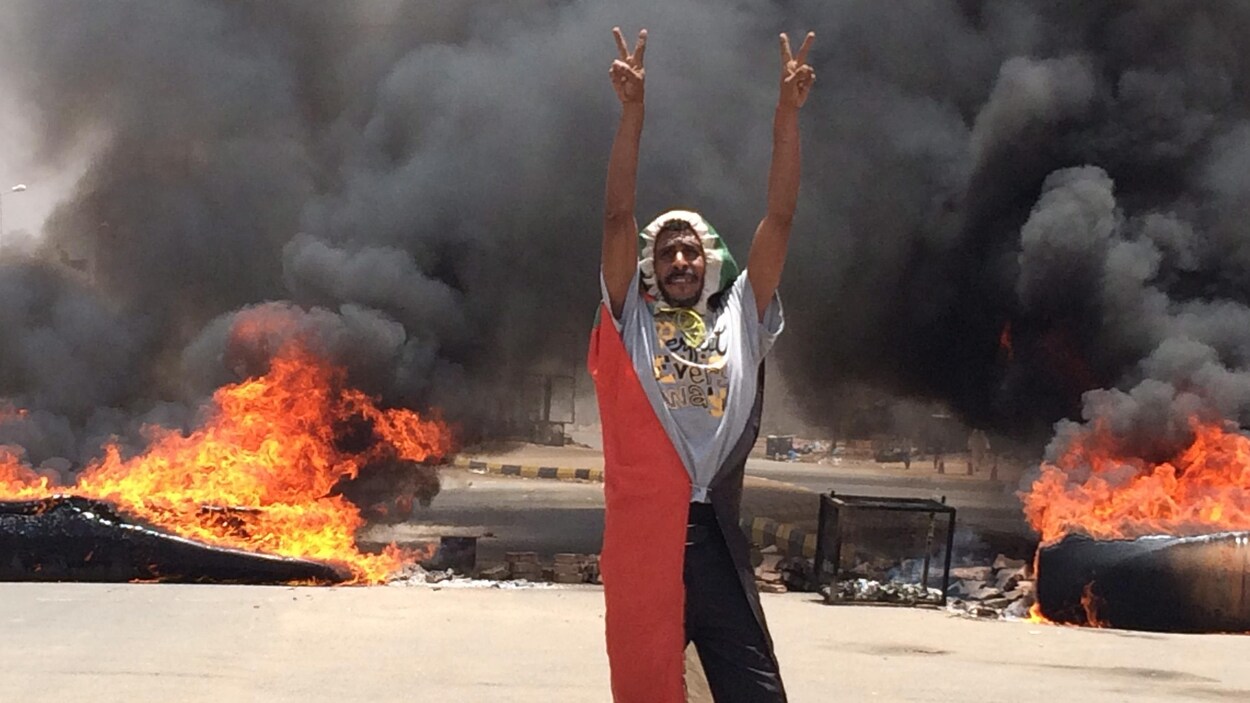 Un homme portant le drapeau soudanais comme une cape fait le signe de la victoire devant des débris en feu.