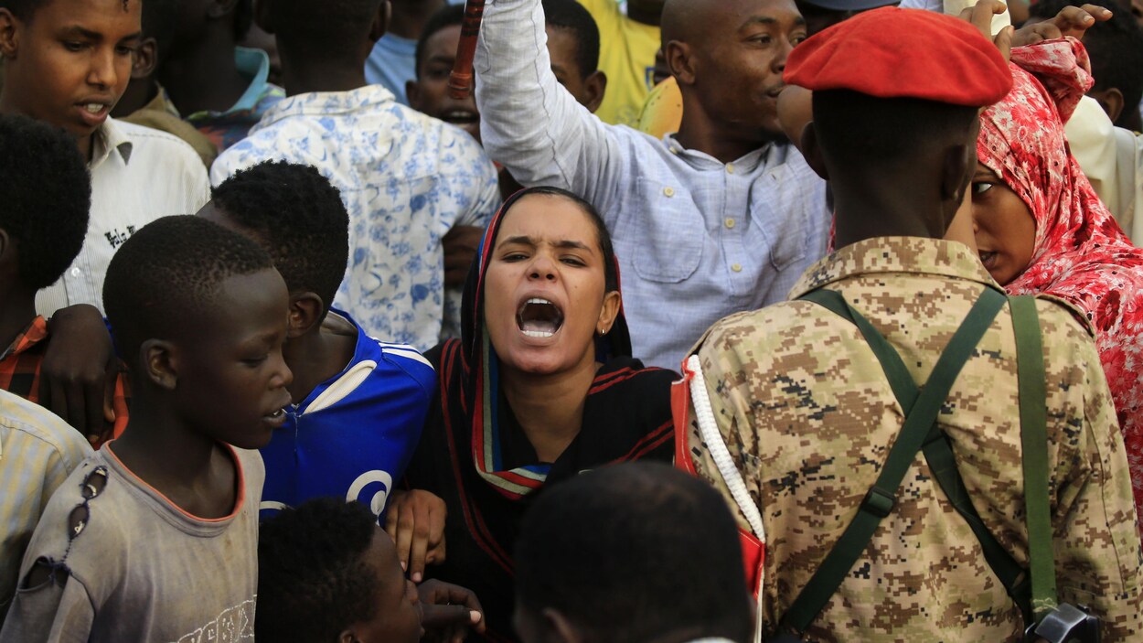 Des femmes, des hommes et des jeunes manifestent dans les rues. Une femme scande un slogan au premier plan, tandis qu'un militaire, de dos, contient la foule.