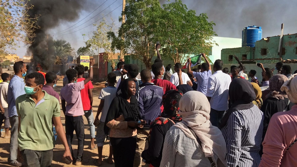 Plusieurs personnes marchent dans une rue de Khartoum. On voit au loin une colonne de fumée noire.
