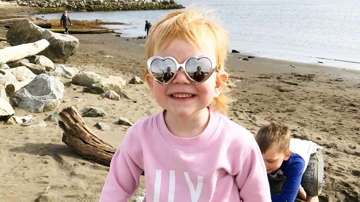 Une petite fille rousse est assise sur un rocher à la plage, porte des lunettes de soleil en forme de coeur et sourit pour l'appareil photo.