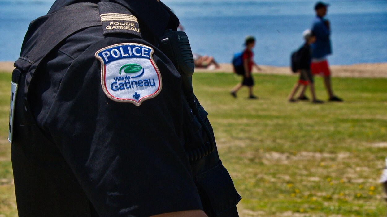 L'épaule d'un policier ayant l'insigne de la police de Gatineau. Des jeunes circulant dans un parc sont visibles en arrière-plan.