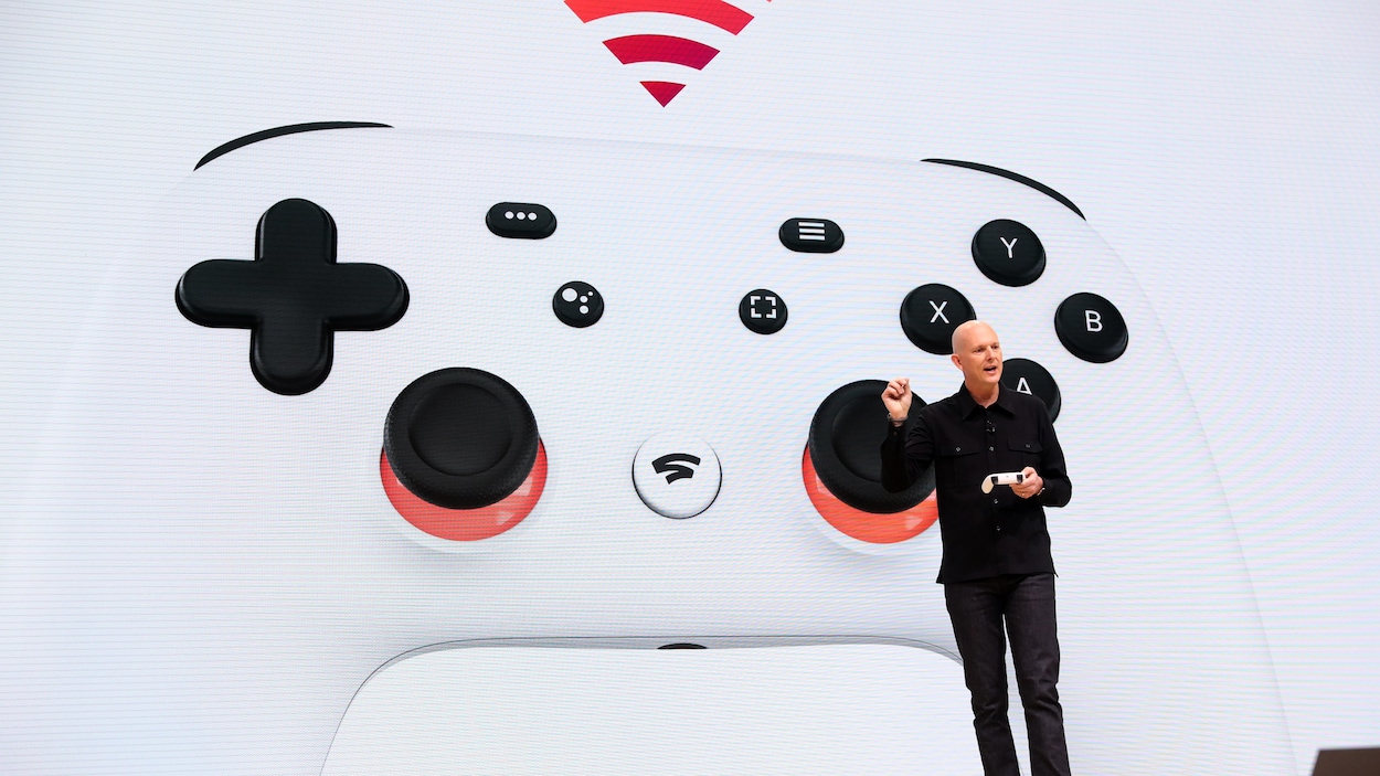 Le vice-président et directeur général de Google Phil Harrison tient la manette du système de jeux vidéo en flux en continu Stadia sur scène, devant une immense projection de celle-ci, lors d'un événement en mars 2019.