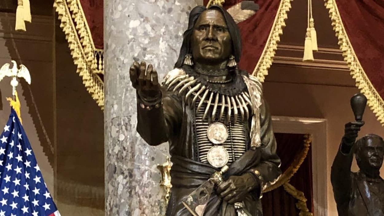 La statue du chef Standing Bear, en tenue de cérémonie, main droite tendue et une hache dans l’autre, à côté du drapeau des États-Unis, dans le grand Hall des statues du Congrès américain.