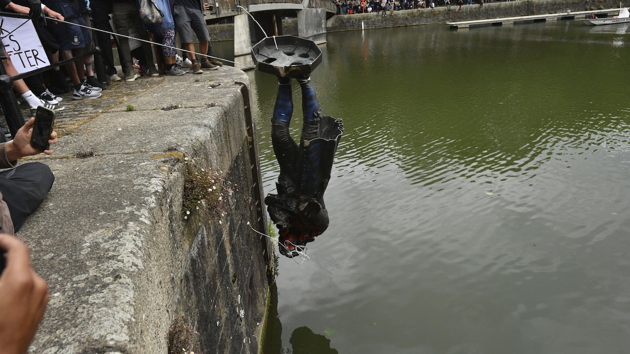Une statue, tête en bas, en retenue par des cordes par des manifestants. Ils s'apprêtent à la jeter dans une rivière.