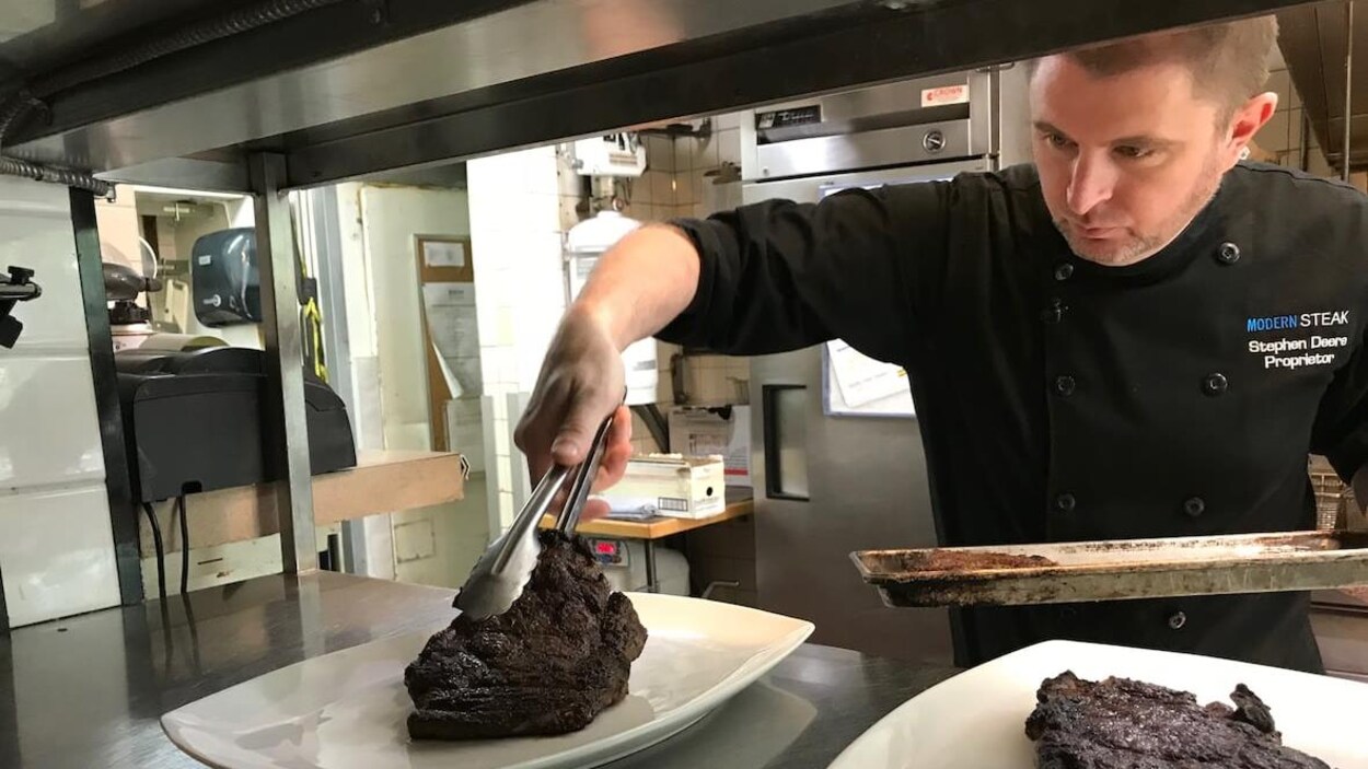 M. Deere, dans la cuisine du restaurant, manipule un gros morceau de boeuf avec des pinces.