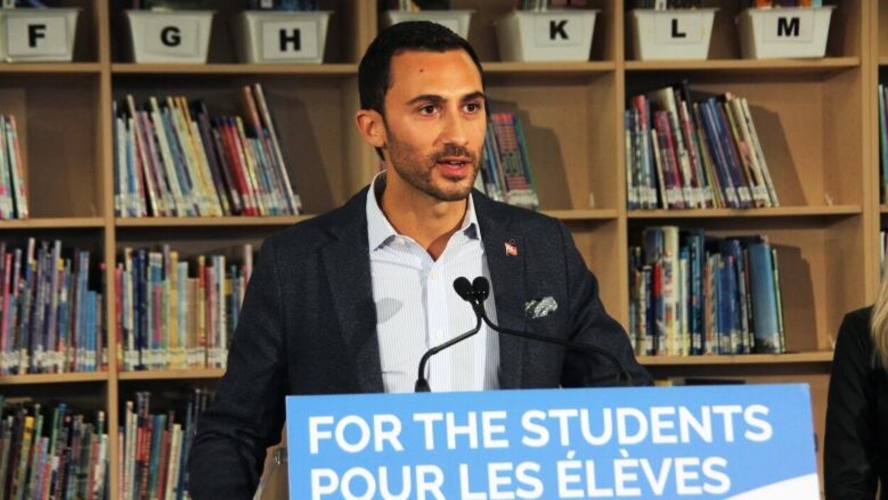 Le ministre de l'Éducation, Stephen Lecce, parle devant un pupitre avec une bibliothèque derrière lui.