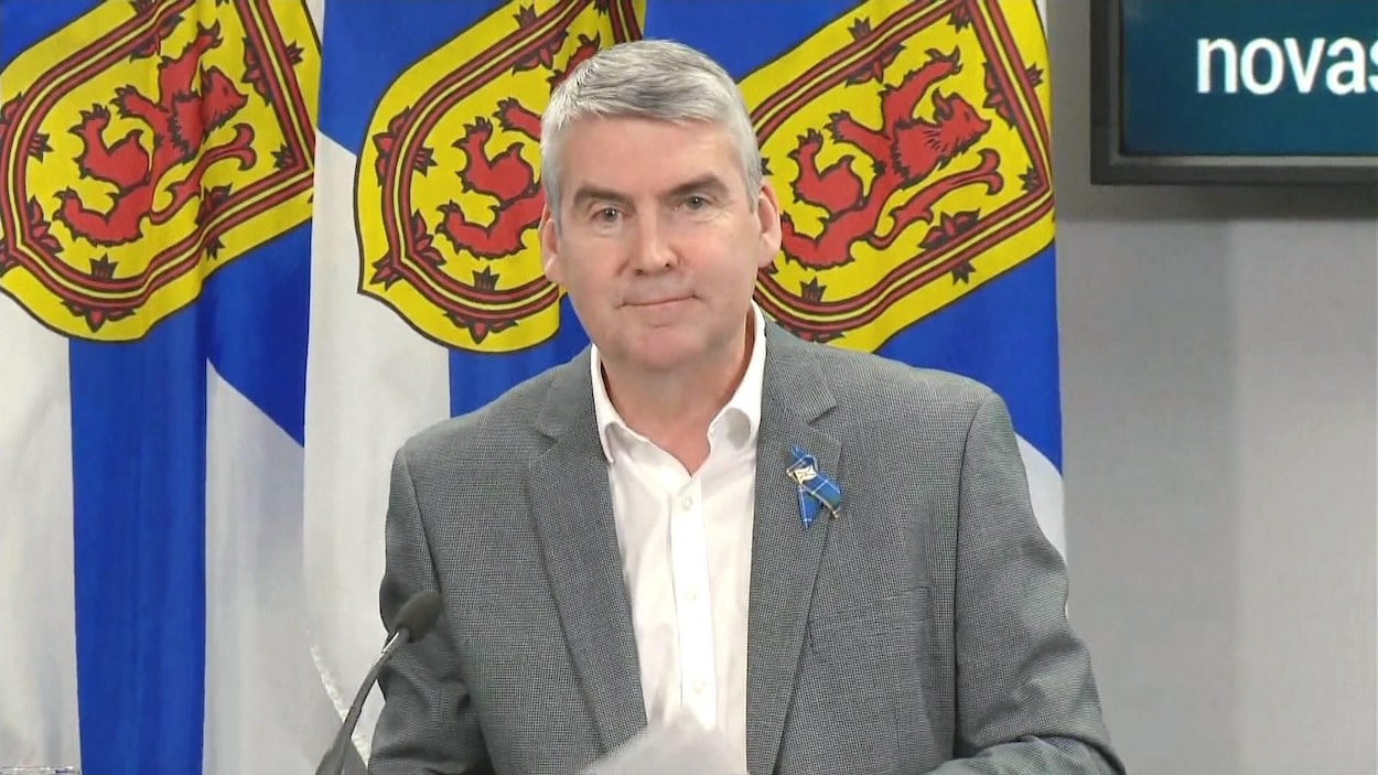 Stephen McNeil, assis et souriant, devant trois drapeaux de la Nouvelle-Écosse.