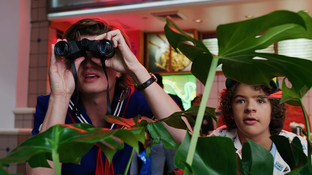 Deux jeunes garçons, dont l'un regarde avec des jumelles, sont cachés derrière une plante.