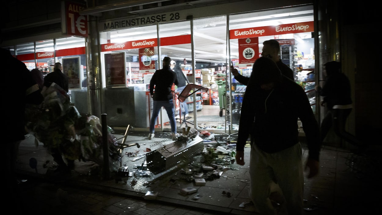 Des produits d'un magasin vandalisé jonchent le sol, dans la nuit, tandis que des gens observent la scène dans la pénombre.