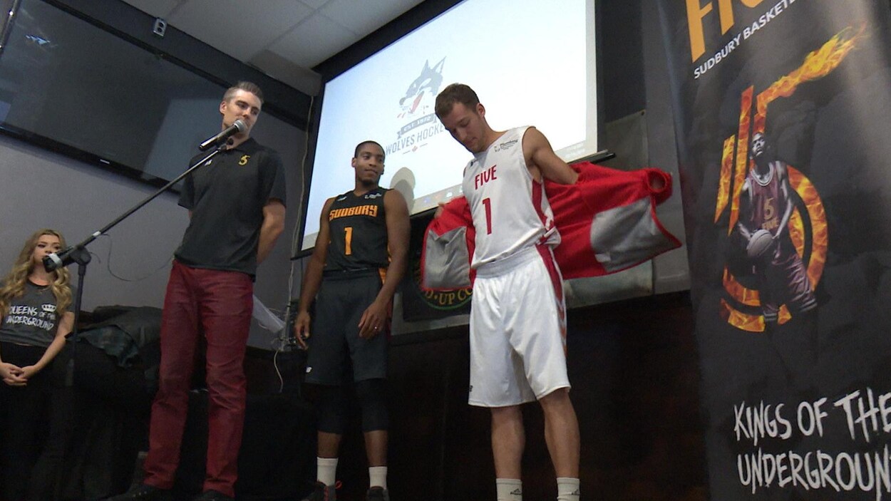 Deux hommes en chandail de basketball se tiennent en compagnie d'un troisième en habit normal.
