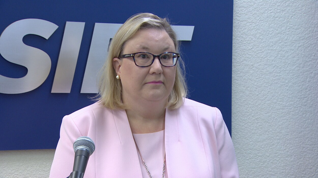 Susan Hughson, avec un veston rose et des lunettes, parle devant un micro avec l'enseigne d'ASIRT derrière elle.