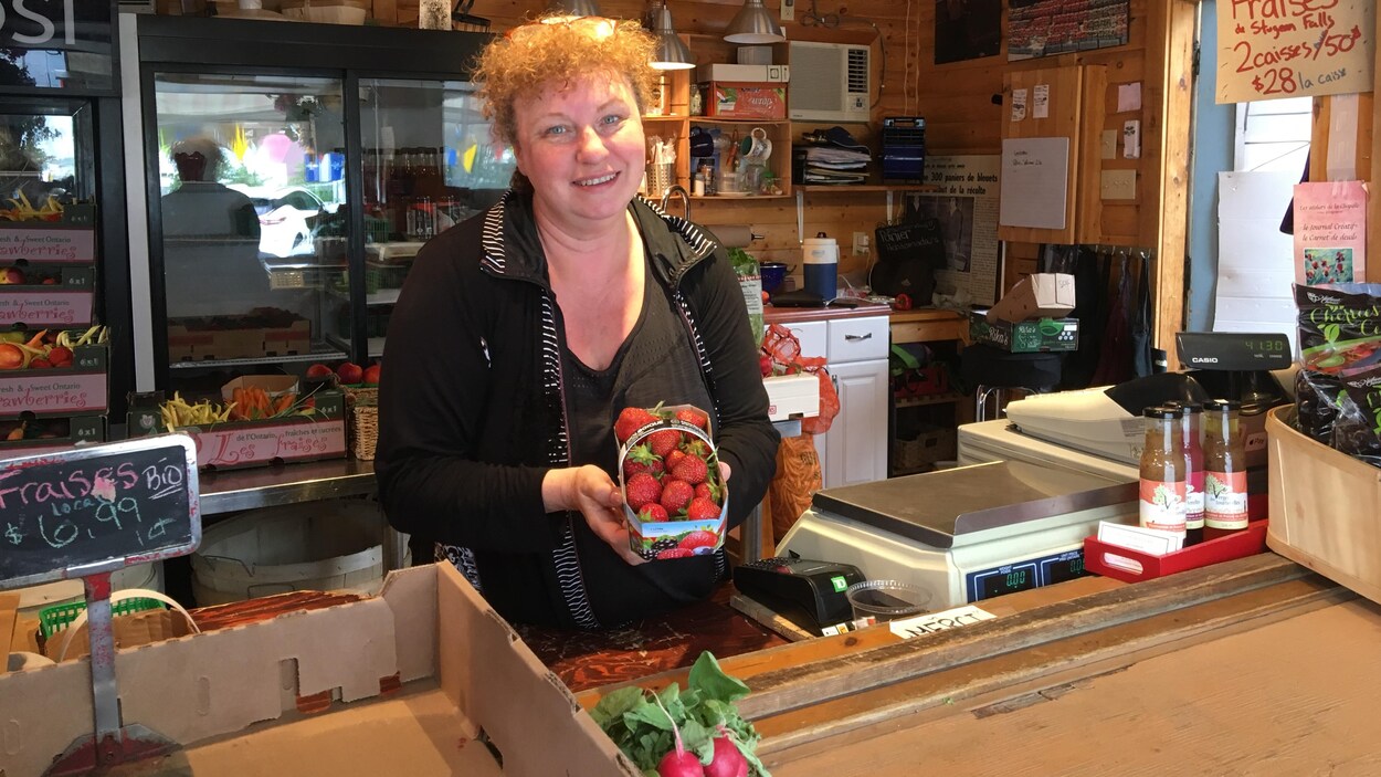 Une femme derrière son comptoir sourit à la caméra en tenant un panier de fraises.