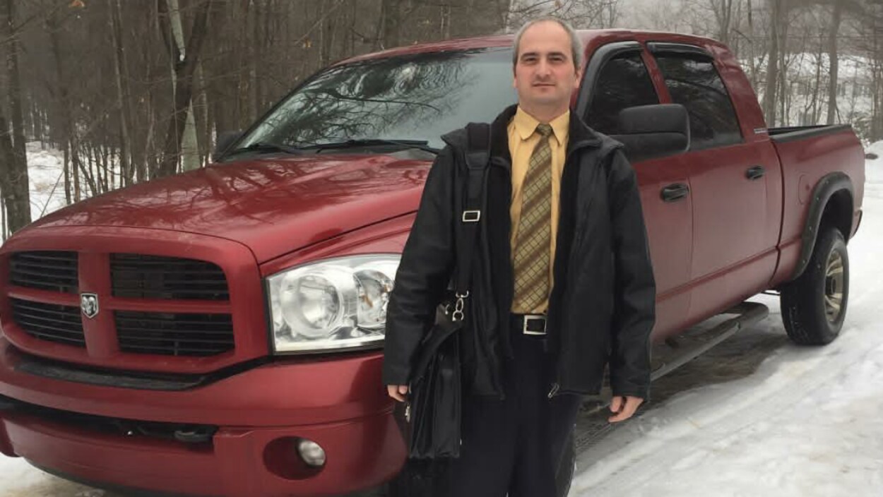 Un homme portant une chemise jaune et un manteau noir pose avec un sac en bandoulière devant une camionnette rouge vin.