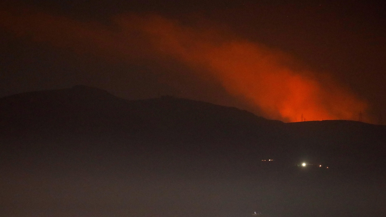 Une vue sur une colonne de fumée rougeâtre s'élevant derrière des montagnes dans la nuit près de Damas, en Syrie.