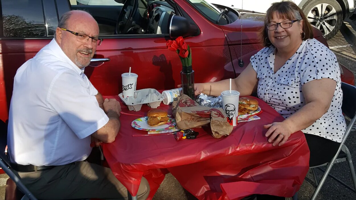 Lisa Hodges et son mari posent pour la caméra. Ils sont dans un stationnement, assis à une table avec des hamburgers de poulet et des boissons gazeuses. Il y a des fleurs dans un vase sur la table.