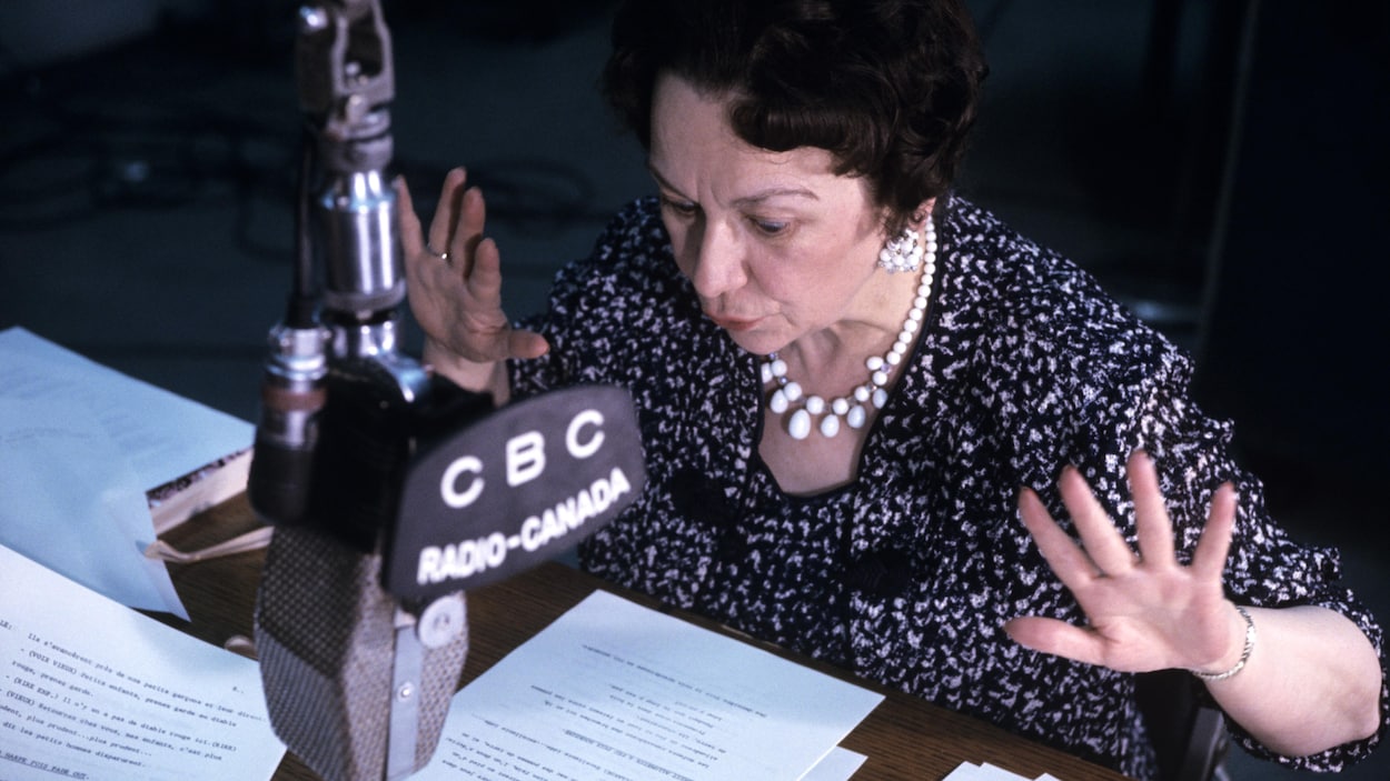 Dans un studio de radio, Lucille Desparois, assise à un pupitre, lit une histoire, les mains en action. Un micro est suspendu devant elle.