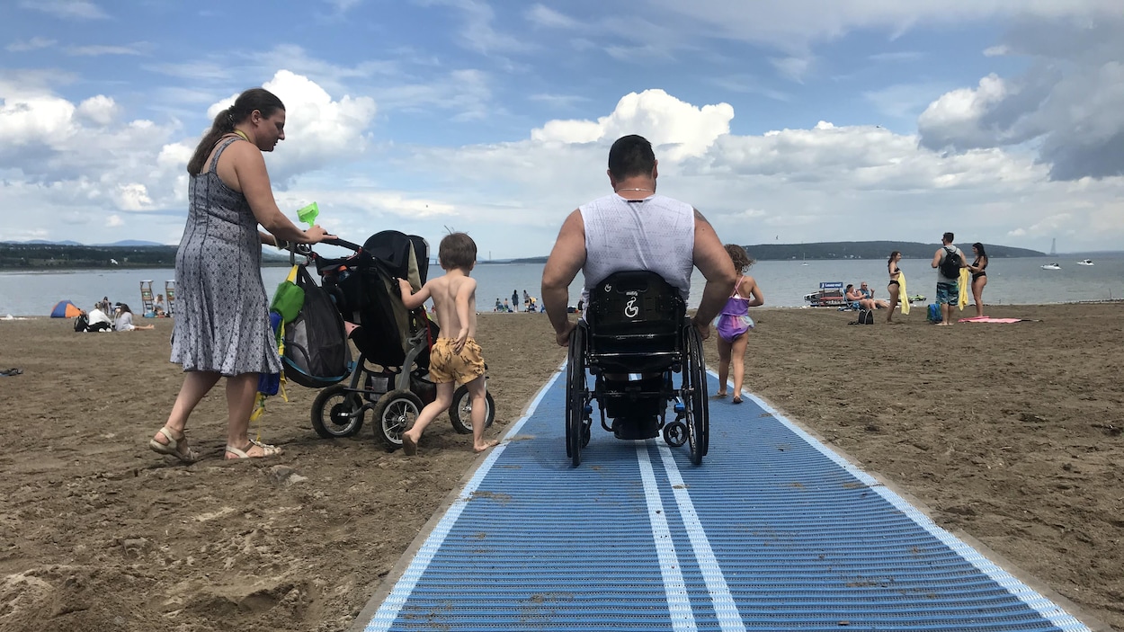 Un homme en fauteuil roulant sur un tapis placé sur le sable d'une plage. Il est avec sa femme et ses enfants.