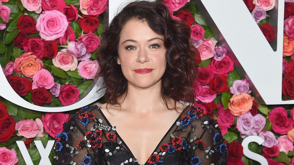 L'actrice canadienne Tatiana Maslany se tient debout devant un mur couvert de roses et sourit.