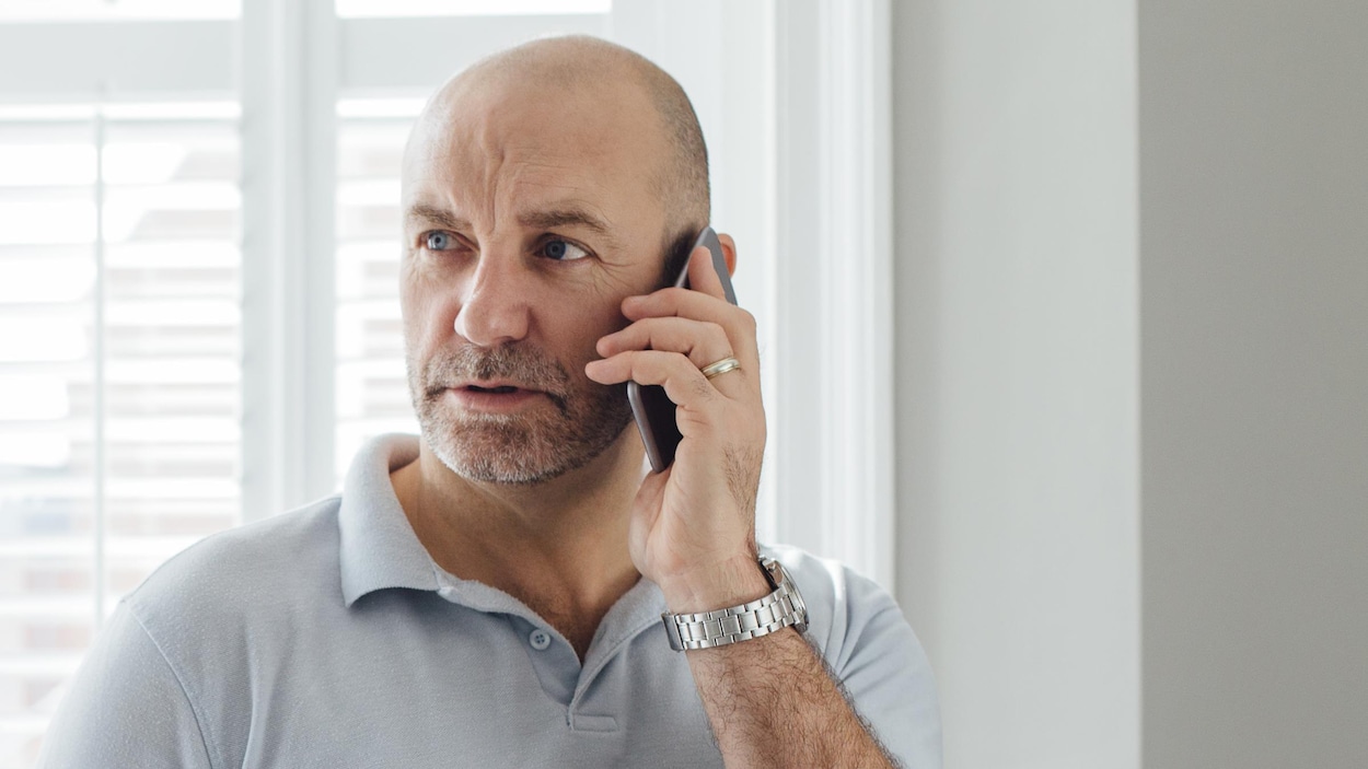 Un homme debout près d'une fenêtre parle au téléphone l'air inquiet.
