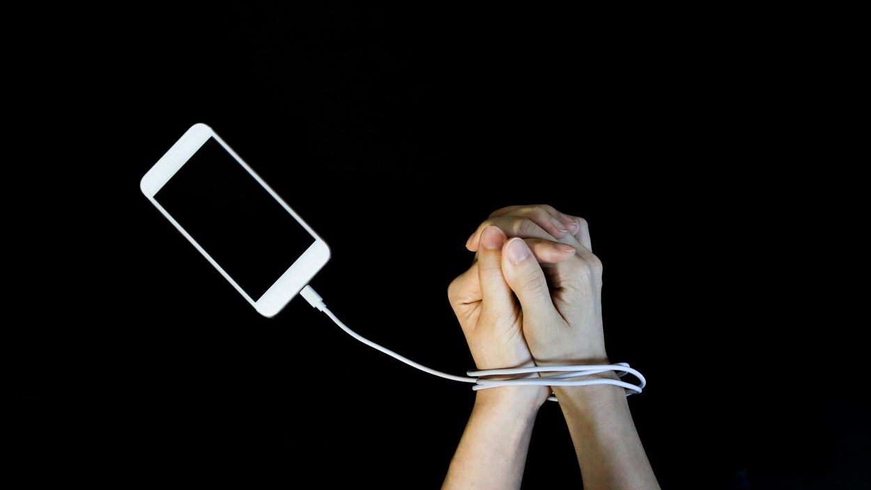 Des mains prises dans un fil de recharge, comme des menottes, branché à un téléphone.