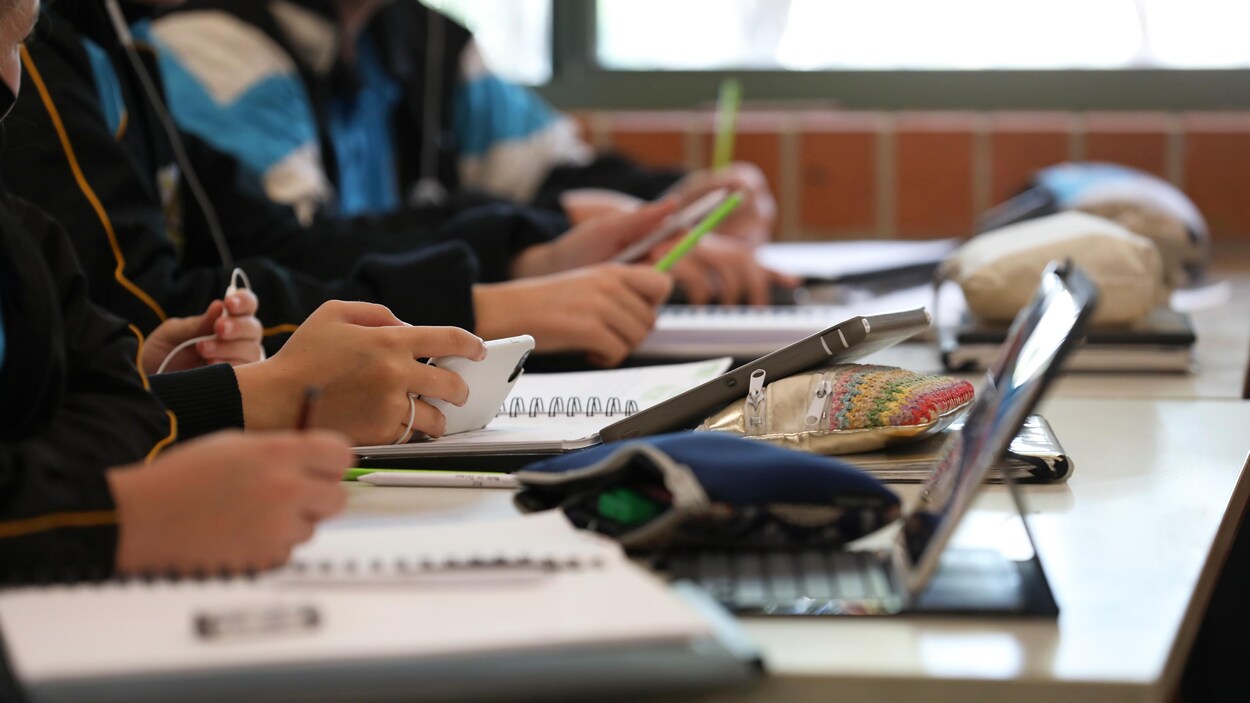 Gros plan de profil sur les mains d'élèves du secondaire qui écrivent dans leurs cahiers ou textent sur leur téléphone, en classe.