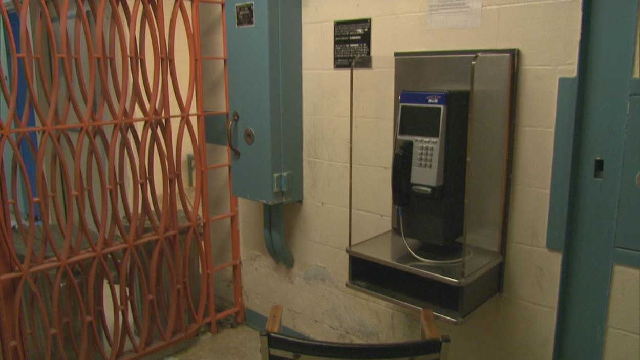 Une chaise est placée devant un téléphone public accroché à un mur de béton. En arrière-plan, une porte en grillage bloque l'accès à un couloir.