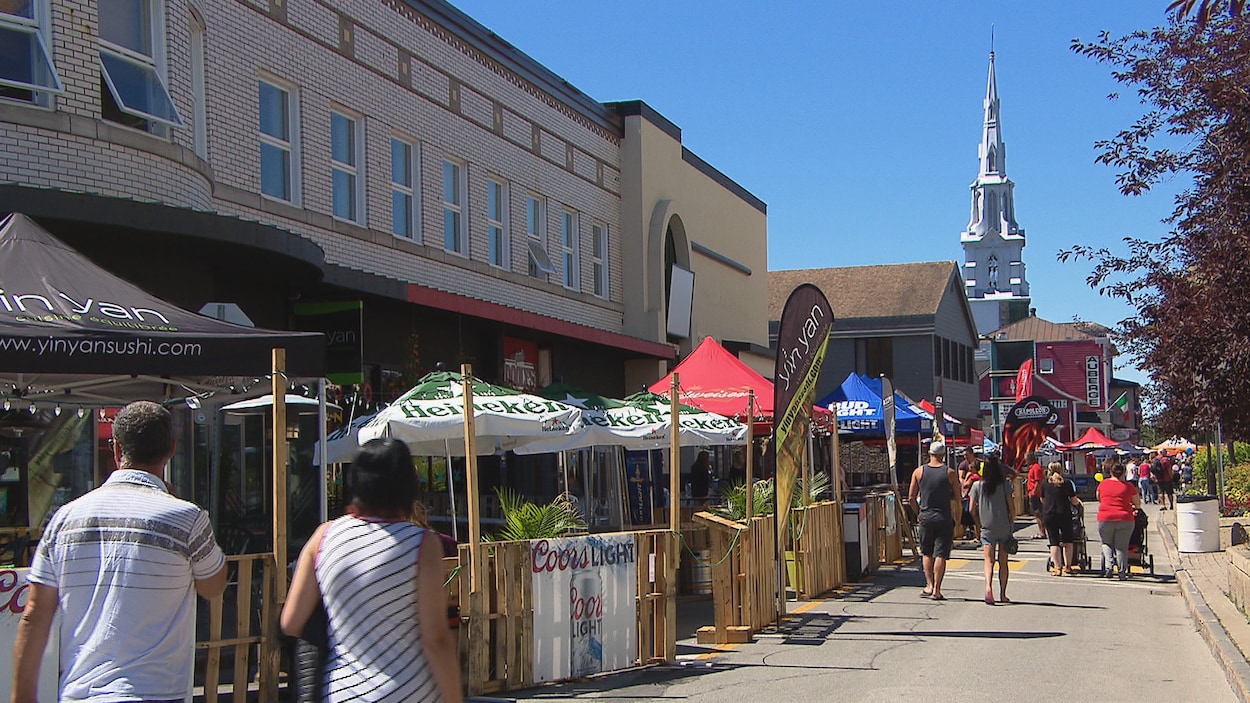 Des gens marchent sur une rue où des terrasses sont installées, chacunes avec leurs parasols.