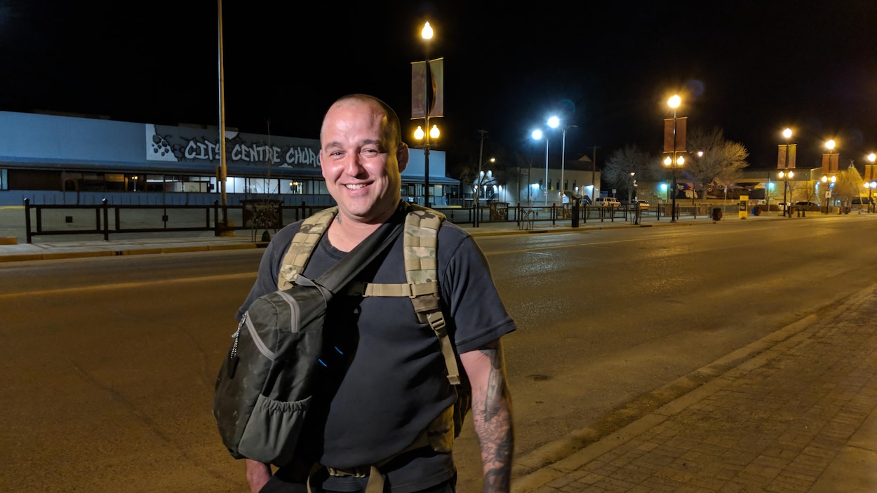 Un homme porte un sac sur son dos et un sac sur sa poitrine dans une rue. Il porte un t-shirt gris et des tatouages. Un homme porte un sac sur son dos et un sac sur sa poitrine dans une rue. Il porte un t-shirt gris et des tatouages.
