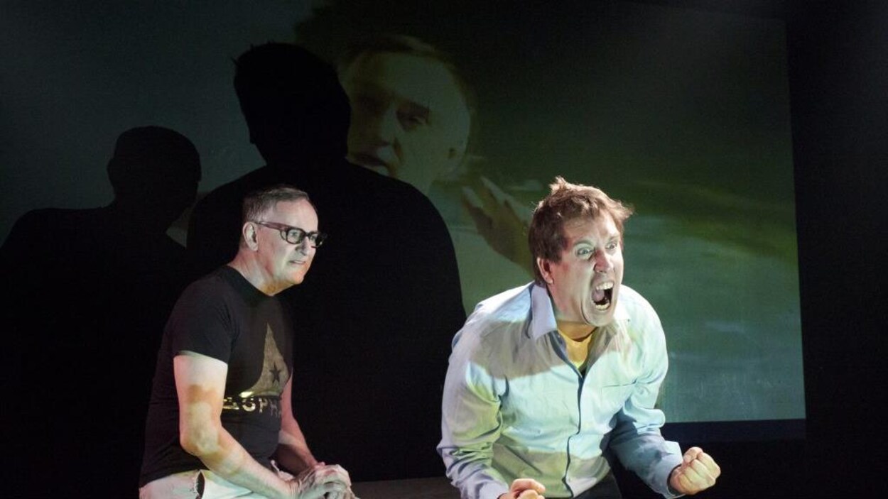Deux hommes sont assis devant un écran où on projette une image de l'un d'eux. L'homme à gauche regarde l'homme à sa droite, qui semble crier de toutes ses forces.