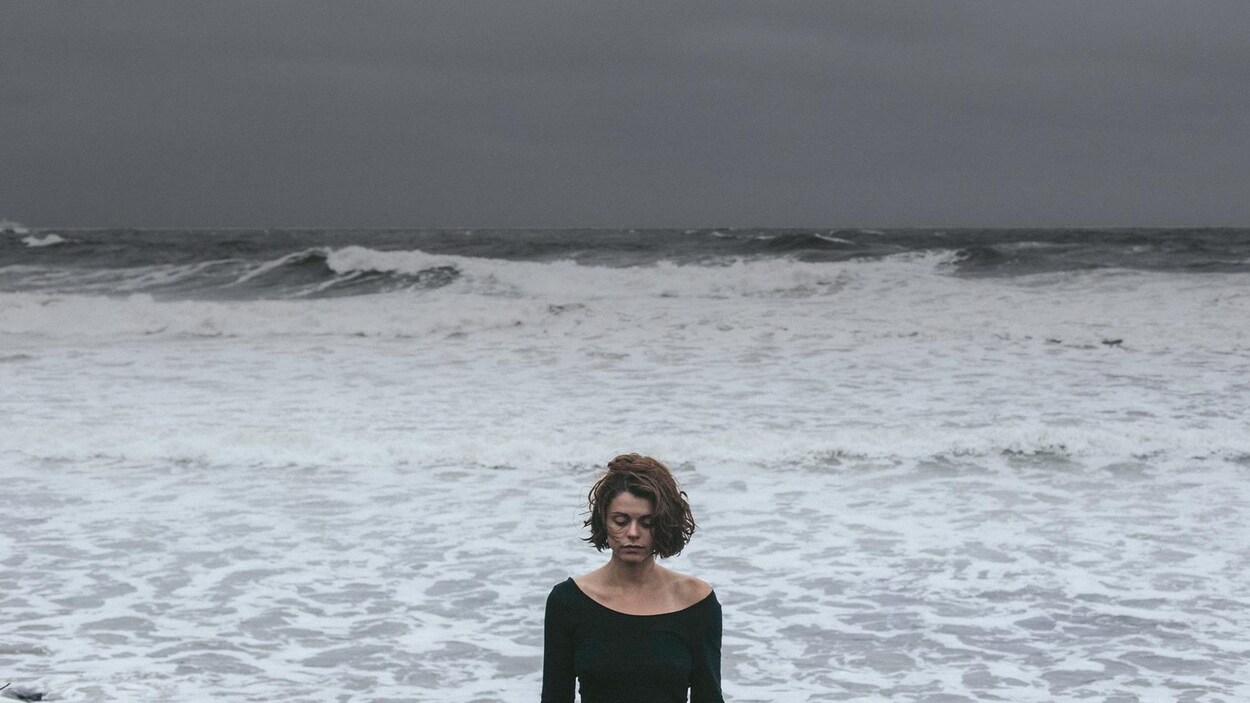 Une femme fait dos aux vagues, au fleuve et au ciel gris