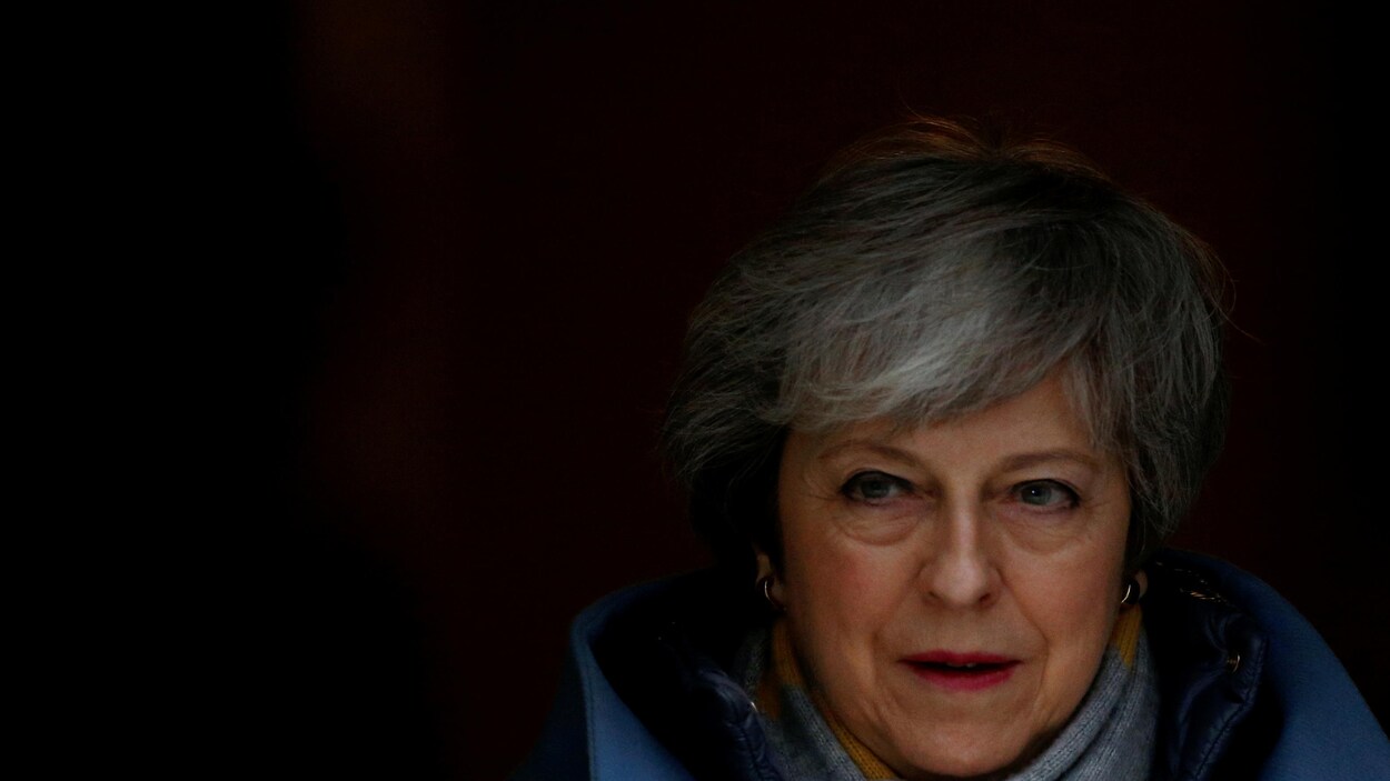 Dans l'ombre, le visage de Theresa May apparaît. Ses yeux verts brillent, ses cheveux sont courts et gris, elle arbore une frange.